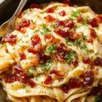 A close-up of a white ceramic plate holding creamy bacon carbonara pasta, topped with crispy bacon bits and freshly grated Parmesan cheese. The pasta is coated in a glossy, rich sauce with visible black pepper specks. The dish is garnished with a sprig of parsley, with a rustic wooden table and lemon wedge in the background adding warmth and contrast.