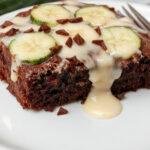 A slice of decadent chocolate zucchini cake with a rich, fudgy texture, topped with a glossy chocolate glaze and garnished with a few zucchini shreds and a sprig of mint. The cake slice is presented on a rustic plate with crumbs scattered around, showcasing the moist interior and smooth frosting contrasts.