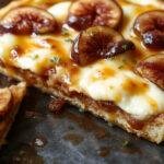 A close-up of a beautifully plated caramelized fig and mascarpone tart. The tart features a golden-brown crust topped with glossy caramelized fig slices arranged in an artistic pattern. The creamy mascarpane layer peeks through, garnished with a dusting of powdered sugar and a sprig of fresh mint. The presentation is elegant, with a rustic wooden table background and soft natural lighting highlighting the textures and colors of the dessert.