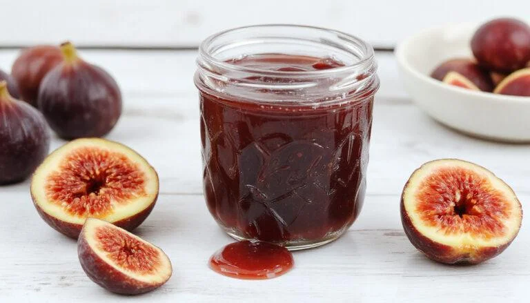 A clear jar of homemade fig jam with a rich amber color, topped with fresh fig slices and a sprig of mint, placed on a rustic wooden surface with a spoon nearby.