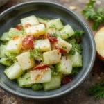 A vibrant, refreshing salad featuring thinly sliced cucumbers, crisp celery sticks, and diced apples arranged on a white ceramic plate. The ingredients are colorful, with the green cucumbers and celery contrasting against the red and yellow apples, dressed lightly with a glossy vinaigrette that gives a subtle sheen. The presentation emphasizes freshness and crunch, with a sprinkle of herbs on top and a backdrop of a rustic wooden table.