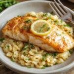 A plated serving of seared salmon on a white ceramic plate, topped with a wedge of lemon. Accompanying the salmon is lemon-colored orzo pasta garnished with fresh herbs, with a drizzle of olive oil around the dish. The salmon has a crispy, golden-brown crust, and the orzo appears fluffy and aromatic, with sprigs of parsley for color contrast.