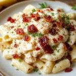 A bowl of creamy crack chicken pasta featuring shredded chicken, crispy bacon pieces, melting cheese, and green herbs, with a golden-brown crust on top, garnished with fresh parsley, presented on a rustic wooden table.