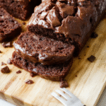 A golden-brown chocolate banana bread loaf slice on a wooden cutting board, glistening with dark chocolate chunks, soft crumb texture, visible banana swirls, dusted with powdered sugar, beside a ripe banana and chocolate shavings on a rustic farmhouse table with natural light.